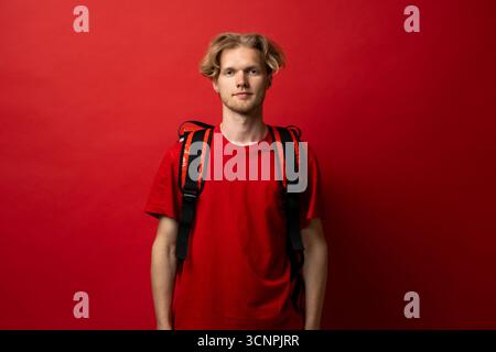 Porträt des jungen Liefermannes in roter Uniform mit Rucksack auf rotem Hintergrund Stockfoto