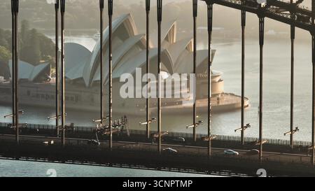 Australien, Sydney: Autos fahren auf der Sydney Harbour Bridge bei Sonnenaufgang, umrahmen das Sydney Opera House im Hintergrund und bieten einen atemberaubenden Blick auf australische Wahrzeichen. Moderne Architektur und städtischer Nahverkehr Stockfoto