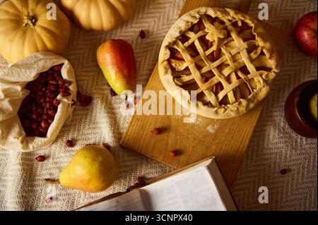 Ein gemütliches Stillleben im Herbst mit einem Gitterkuchen auf einem Holzbrett, Kürbissen, Äpfeln, Birnen und getrockneten Preiselbeeren in einer Tasche. Ein geöffnetes Buch schließt das ab Stockfoto