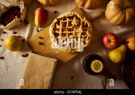 Eine gemütliche Herbstszene zeigt einen Kuchen mit Gitteroberseite auf einem Holzbrett, umgeben von Kürbissen, Äpfeln, Birnen, einer Zitrone im Becher und Preiselbeeren. Warm, Rost Stockfoto