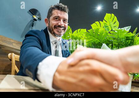 Ein selbstbewusster Geschäftsmann im Anzug lächelt warm und schüttelt die Hände über einen Schreibtisch Stockfoto