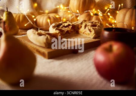 Eine gemütliche Herbstszene mit Apfel- und Kürbiskuchen auf einem hölzernen Schneidebrett, umgeben von Kürbissen, Birnen und warmen Lichtern, die Thank festhalten Stockfoto