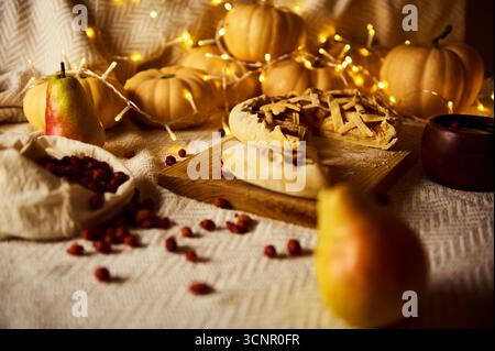 Gemütliches Stillleben im Herbst mit Kuchen auf einem Holzbrett, Birnen, Preiselbeeren, Kürbissen und warmen Lichtern, die eine gemütliche Thanksgiving-Stimmung hervorrufen. Stockfoto