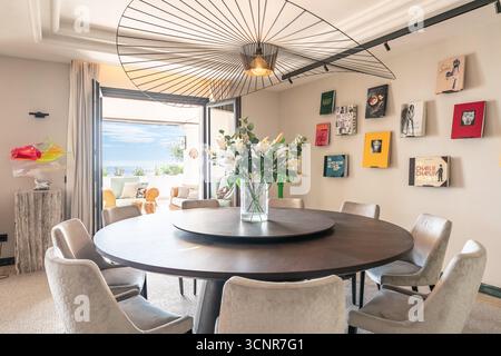 Elegantes, modernes Esszimmer mit großem runden Holztisch mit Polsterstühlen, einem skulpturalen Designer-Kronleuchter und einer Glasvase Stockfoto