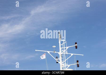 Ein Blick aus der Nähe auf einen weißen Schiffsmast mit Navigationslichtern und Takelage vor einem hellblauen Himmel, der nautische Technologie, Sicherheit und maritim vermittelt Stockfoto