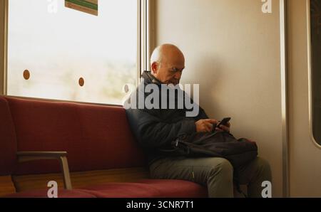 Ein älterer Mann, der sich während einer ruhigen Zugfahrt durch die Landschaft von Nara in sein Handy verschlungen hat Stockfoto
