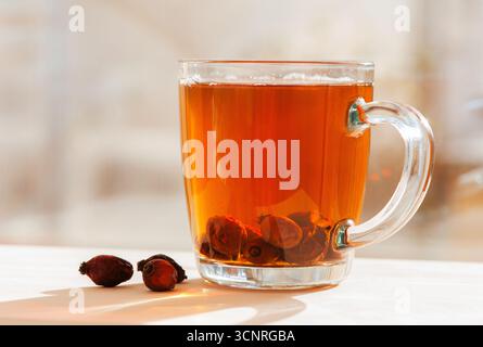 Eine Glasschale Hagebuttentee mit getrockneten Beeren innen und auf dem Tisch bei warmem Sonnenlicht verstreut. Stockfoto