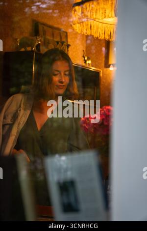 Eine Frau in einem grünen Kleid wird durch ein Fenster in einem gemütlichen Vintage-Zimmer mit warmer Beleuchtung und einem Blumenstrauß gesehen. Peredelkino Stockfoto