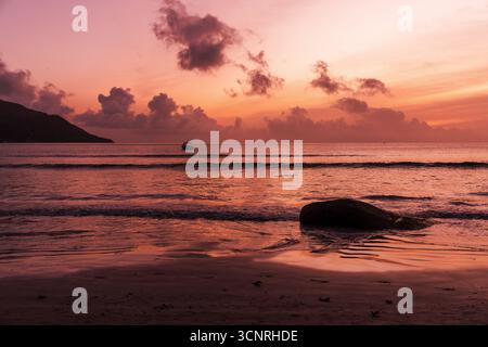Eine malerische Strandszene mit glatten Felsen und ruhigem Wasser, beleuchtet von oramgenrotem Sonnenuntergang. Küstenlandschaft der Insel Mahe, Seychellen Stockfoto