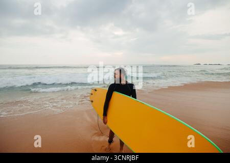 Junger bärtiger Mann mit Surfbrett, der in der Nähe eines Strandes steht. Mann mit Surfbrett im Freien an einem Sommertag. Stockfoto
