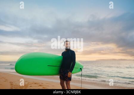 Junger bärtiger Mann mit Surfbrett, der in der Nähe eines Strandes steht. Mann mit Surfbrett im Freien an einem Sommertag. Stockfoto