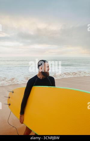 Junger bärtiger Mann mit Surfbrett, der in der Nähe eines Strandes steht. Mann mit Surfbrett im Freien an einem Sommertag. Stockfoto