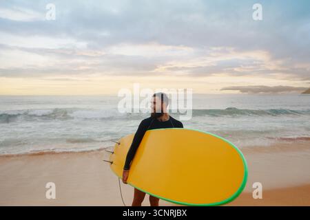Junger bärtiger Mann mit Surfbrett, der in der Nähe eines Strandes steht. Mann mit Surfbrett im Freien an einem Sommertag. Stockfoto