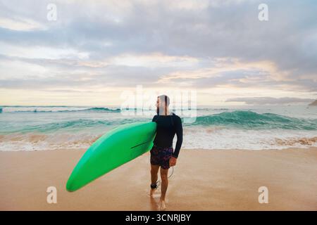 Junger bärtiger Mann mit Surfbrett, der in der Nähe eines Strandes steht. Mann mit Surfbrett im Freien an einem Sommertag. Stockfoto