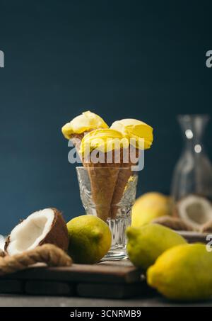 Eine vertikale Nahaufnahme von drei zitronengelben Eiskegeln in einem Glas, umgeben von Zitronen und einer halben Kokosnuss Stockfoto