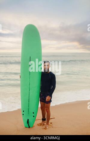 Junger bärtiger Mann mit Surfbrett, der in der Nähe eines Strandes steht. Mann mit Surfbrett im Freien an einem Sommertag. Stockfoto