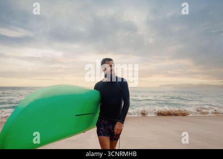 Junger bärtiger Mann mit Surfbrett, der in der Nähe eines Strandes steht. Mann mit Surfbrett im Freien an einem Sommertag. Stockfoto