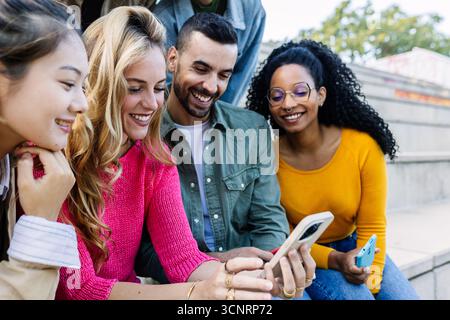 Glückliche multirassische Millennial-Freunde, die sich im Freien mit Mobiltelefonen amüsieren Stockfoto
