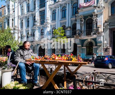 Kiew, Ukraine 11. Mai 2025, Straßenverkäufer, der an einem sonnigen Tag in Zentral-Kiew, Ukraine, lebhaftes handgefertigtes Kunsthandwerk anbietet, mit kunstvoller historischer Gebäudefassade und urbanem Leben im Hintergrund Stockfoto
