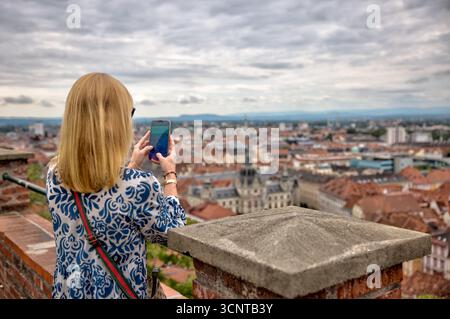 Eine Frau fotografiert den Panoramablick auf das historische Stadtzentrum mit roten Dächern, Kirchtürmen und üppigem Sommerlaub Stockfoto