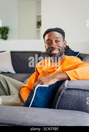 Afroamerikanischer Mann, der sich auf der grauen Couch im Wohnzimmer entspannt, mit orangefarbenem Sweatshirt und Pflanze Stockfoto