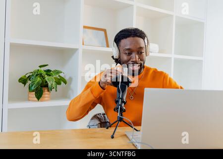 Afroamerikanischer Mann am Schreibtisch, der Pullover-Kopfhörer trägt, der in das Mikrofon spricht, mit einem Becher am Laptop Stockfoto