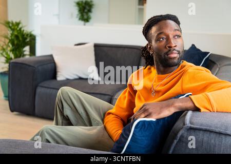 Afroamerikaner sitzt zu Hause auf dem Sofa und trägt ein orangefarbenes Sweatshirt neben Topfpflanzen Stockfoto