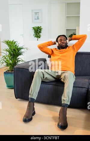Afroamerikanischer Erwachsener, der sich mit kabellosen Kopfhörern auf dem dunkelgrauen Sofa im Wohnzimmer zurücklehnt Stockfoto