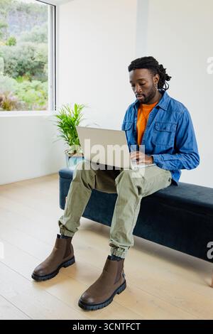 Afroamerikaner tippt auf silbernem Laptop, sitzt auf einer Bank mit Topfpflanzen in hellem Raum Stockfoto