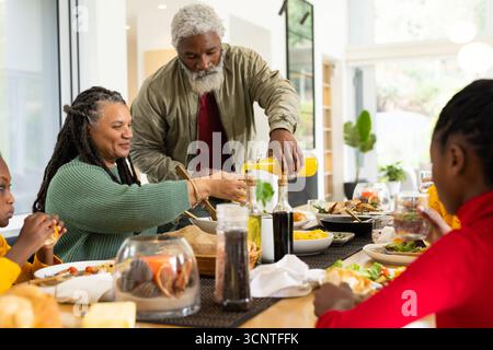 Eine vielfältige Familie, die zu Hause um einen hölzernen Esstisch sitzt und Orangensaft und Salat serviert Stockfoto