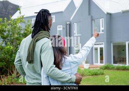 Verschiedene Paare stehen auf grasbewachsenem Rasen vor grauem zweistöckigem Haus, das in Richtung Berghang zeigt Stockfoto