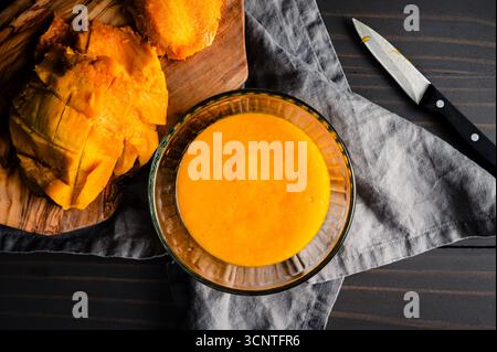 Hausgemachtes Mangopüree in einer Glasschüssel: Pürierte tropische Früchte mit Mangohälte und Samen auf einem hölzernen Schneidebrett Stockfoto