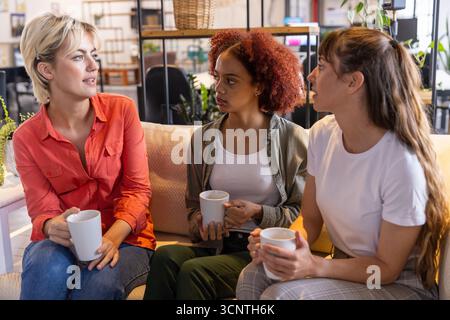 Verschiedene weibliche Kollegen sitzen auf der Couch in der Bürolounge und schlürfen Kaffee aus Keramikbechern Stockfoto