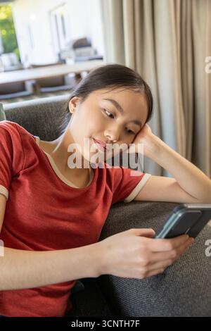 Asiatische Frau, die zu Hause auf dem Sofa am Fenster liegt und Smartphone hält, dekoratives Kissen Stockfoto