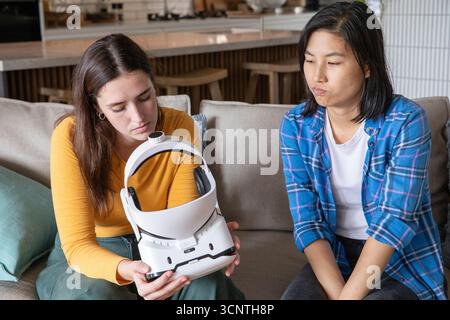 Verschiedene Freundinnen untersuchen das weiße Headset, während sie in der modernen Küche auf der beigefarbenen Couch sitzen Stockfoto