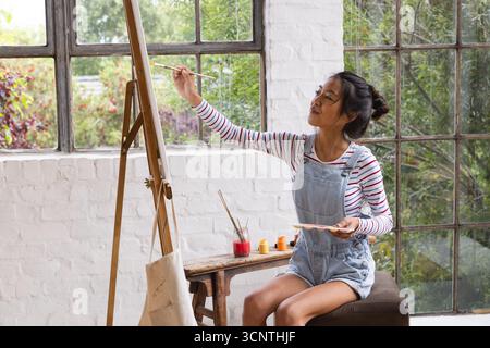 Asiatische Malerin, die auf dem Stuhl sitzt und Leinwand auf Staffelei im Atelier mit Palette und Pinsel malte Stockfoto