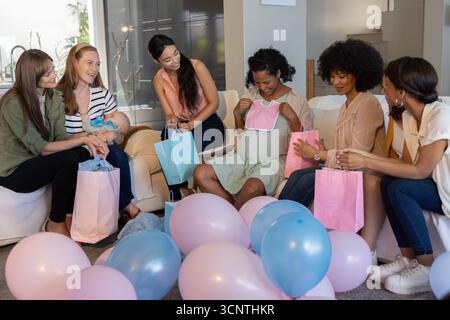Diverse Freundinnen feiern Babydusche im Wohnzimmer, Pastellballons Geschenktüten Lätzchen Stockfoto