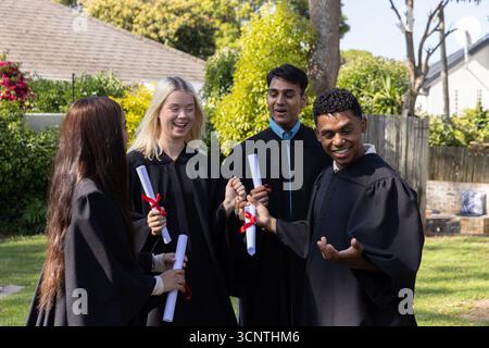 Verschiedene Absolventen in schwarzen Gewändern und mit gerollten Bändchen-Diplomen, die im Hinterhof lachen Stockfoto