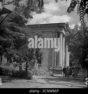 Ein eindrucksvolles Schwarz-weiß-Archivfoto aus den späten 1950er Jahren zeigt die beeindruckende neoklassizistische Architektur der Pädagogischen Universität in Sloviansk, Ukrainische SSR. Das Bild konzentriert sich auf einen großen Säulengang mit hoch aufragenden Säulen, während sich der Alltag um ihn herum entfaltet, mit Fußgängern, die auf dem Bürgersteig spazieren und einer Frau, die mit dem Fahrrad steht. Diese Ansicht fängt die Integration monumentaler sowjetischer Bildungseinrichtungen in das tägliche Gefüge einer Provinzstadt ein Stockfoto