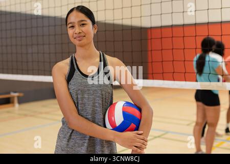 Verschiedene weibliche Mannschaftskameraden stehen am Netz und halten rot-weiß-blaues Volleyball mit Bank im Fitnessstudio Stockfoto