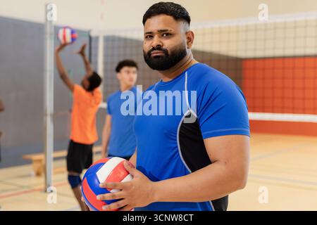 Verschiedene männliche Mannschaftskameraden in Sportbekleidung, die Volleyball halten, im Fitnessstudio in der Nähe des Netzes springen Stockfoto