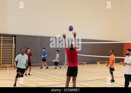 Verschiedene männliche Freunde spielen lässiges Volleyballspiel im Turnsaal mit Netz und in Sportbekleidung Stockfoto