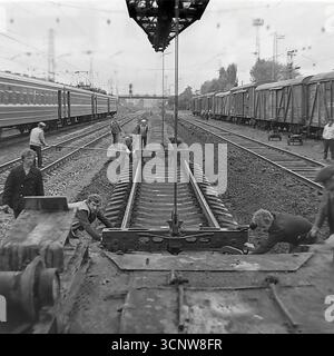 Ein dynamisches Archivfoto aus den 1970er Jahren zeigt ein Team sowjetischer Eisenbahner, die in Sloviansk, der ukrainischen SSR, mit schweren Arbeiten am Gleisbau beschäftigt waren. Mit Hilfe eines speziellen Gleiskrans manövriert und installiert die Besatzung gemeinsam einen großen, vorgefertigten Abschnitt der Eisenbahnstrecke mit Betonschwellen. Dieses leistungsstarke Schwarzweiß-Bild dokumentiert einen Schlüsselmoment in einem großen Infrastrukturprojekt und unterstreicht die Synergie von Handarbeit und schweren Maschinen in der UdSSR Stockfoto