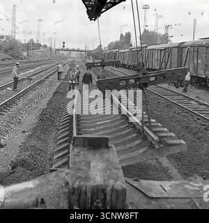 Ein dynamisches Archivfoto aus den 1970er Jahren zeigt ein Team sowjetischer Eisenbahner, die in Sloviansk, der ukrainischen SSR, mit schweren Arbeiten am Gleisbau beschäftigt waren. Mit Hilfe eines speziellen Gleiskrans manövriert und installiert die Besatzung gemeinsam einen großen, vorgefertigten Abschnitt der Eisenbahnstrecke mit Betonschwellen. Dieses leistungsstarke Schwarzweiß-Bild dokumentiert einen Schlüsselmoment in einem großen Infrastrukturprojekt und unterstreicht die Synergie von Handarbeit und schweren Maschinen in der UdSSR Stockfoto