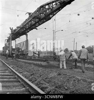 Ein dynamisches Archivfoto aus den 1970er Jahren zeigt ein sowjetisches Eisenbahnbaupersonal in Sloviansk, der ukrainischen SSR. Das Team von Arbeitern arbeitet zusammen, um einen schweren, vorgefertigten Abschnitt der Schiene in Position zu führen, während er von einem massiven Portalkran abgesenkt wird. Dieses Schwarzweiß-Bild zeigt anschaulich die Synergie zwischen Handarbeit und schweren Maschinen während eines Gleisinstallationsprojekts auf einer elektrifizierten Eisenbahnstrecke und zeigt einen Moment intensiver, koordinierter Anstrengungen Stockfoto