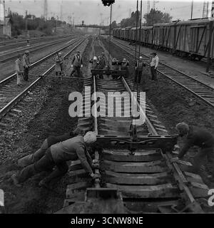Ein dynamisches Archivfoto aus den 1970er Jahren zeigt ein Team sowjetischer Eisenbahner, die in Sloviansk, der ukrainischen SSR, mit schweren Arbeiten am Gleisbau beschäftigt waren. Mit Hilfe eines speziellen Gleiskrans manövriert und installiert die Besatzung gemeinsam einen großen, vorgefertigten Abschnitt der Eisenbahnstrecke mit Betonschwellen. Dieses leistungsstarke Schwarzweiß-Bild dokumentiert einen Schlüsselmoment in einem großen Infrastrukturprojekt und unterstreicht die Synergie von Handarbeit und schweren Maschinen in der UdSSR Stockfoto