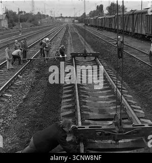 Ein dynamisches Archivfoto aus den 1970er Jahren zeigt ein Team sowjetischer Eisenbahner, die in Sloviansk, der ukrainischen SSR, mit schweren Arbeiten am Gleisbau beschäftigt waren. Mit Hilfe eines speziellen Gleiskrans manövriert und installiert die Besatzung gemeinsam einen großen, vorgefertigten Abschnitt der Eisenbahnstrecke mit Betonschwellen. Dieses leistungsstarke Schwarzweiß-Bild dokumentiert einen Schlüsselmoment in einem großen Infrastrukturprojekt und unterstreicht die Synergie von Handarbeit und schweren Maschinen in der UdSSR Stockfoto