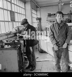 Ein atmosphärisches Archivfoto aus den 1980er Jahren zeigt das Innere einer sowjetischen Autowerkstatt in Sloviansk, ukrainische SSR. Zwei Mechaniker sind in ihrer typischen Arbeitsumgebung zu sehen: Ein Mann konzentriert sich auf eine Aufgabe an seiner Werkbank an den großen Fenstern, während sein jüngerer Kollege inne hält, um für die Kamera zu posieren und einen Schraubenschlüssel zu halten. Dieses ehrliche Schwarzweiß-Bild bietet ein authentisches Stück Leben und zeigt die Werkzeuge, Maschinen und die tägliche Routine eines Arbeiterberufs in der UdSSR Stockfoto