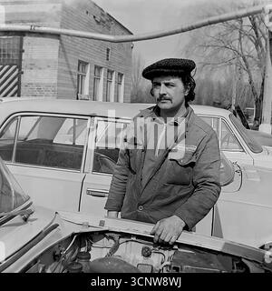 Ein fesselndes und charakterreiches Archivporträt aus den 1980er Jahren zeigt einen sowjetischen Automechaniker oder Fahrer in Sloviansk, der ukrainischen SSR. Der gutaussehende Mann mit Schnurrbart und stylischer flacher Kappe steht selbstbewusst neben einem VAZ „Zhiguli“-Auto mit offener Motorhaube und offenbart den Motor. Dieses Schwarzweiß-Foto fängt die ruhige würde und den professionellen Stolz der sowjetischen Arbeiterklasse ein und bietet einen authentischen Einblick in die Atmosphäre eines Fahrzeugreparaturhofs während dieser Zeit Stockfoto
