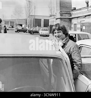 Ein wunderbar charismatisches und freundliches Archivporträt aus den 1980er Jahren zeigt einen hübschen jungen sowjetischen Fahrer oder Arbeiter mit einem charmanten Lächeln und einem stilvollen Haarschnitt. In einem großen Fahrzeugdepot in Sloviansk, der ukrainischen SSR, neben seinem Auto stehend, fängt der warme und einnehmende Ausdruck des Mannes den optimistischen Geist der Jugend in der späten sowjetischen Ära ein. Dieses authentische Schwarzweiß-Foto ist ein seltener, positiver Einblick in den Alltag und die Persönlichkeit eines jungen Arbeiters Stockfoto